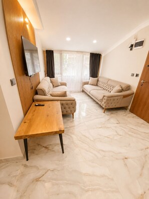 A modern living room with a wooden table, two sofas, a flat-screen TV mounted on a wooden panel, and a large window with curtains. - Sefa Suit (Caykara)