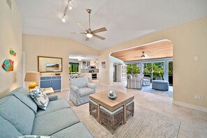 Uma sala de estar ampla com sofá, poltronas e uma mesa de centro. Uma cozinha aparece ao fundo e há uma varanda com espaço para sentar ao ar livre.