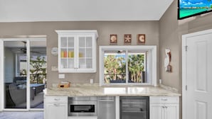 A kitchen with white cabinetry, a built-in microwave, and a dishwasher. There are two windows with views of palm trees and a balcony. - Paradise Point: Beautiful Pool Home in Beachview! (Sanibel)