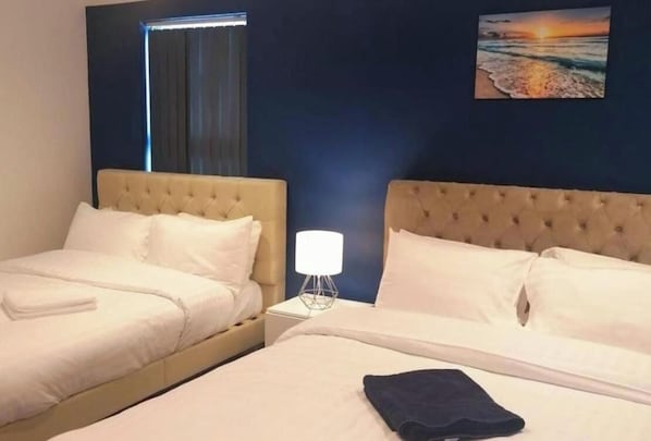 A hotel room with two beds, a nightstand with a lamp, a painting of a sunset, and a window with curtains.
