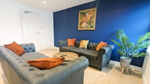 Ein Wohnzimmer mit dunkelblauer Akzentwand, einer grauen Couch mit orangen Kissen, einem Holztisch und einer Topfpflanze.