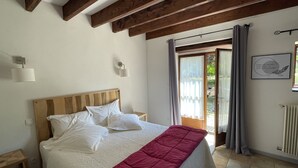 Un dormitorio con cabecera de madera, cama con ropa de cama blanca, manta roja, mesita de noche, lámpara de pared, ventana con cortinas y una cita enmarcada en la pared.
