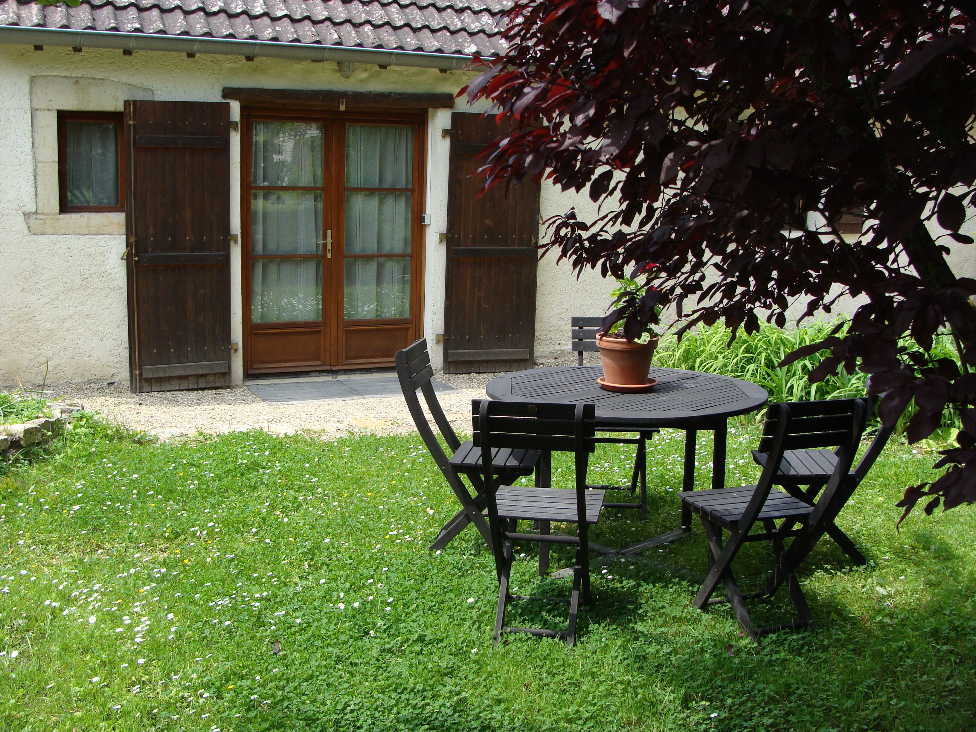 A small outdoor seating area with a round table and four chairs, a potted plant, and a house with a tiled roof and wooden doors.