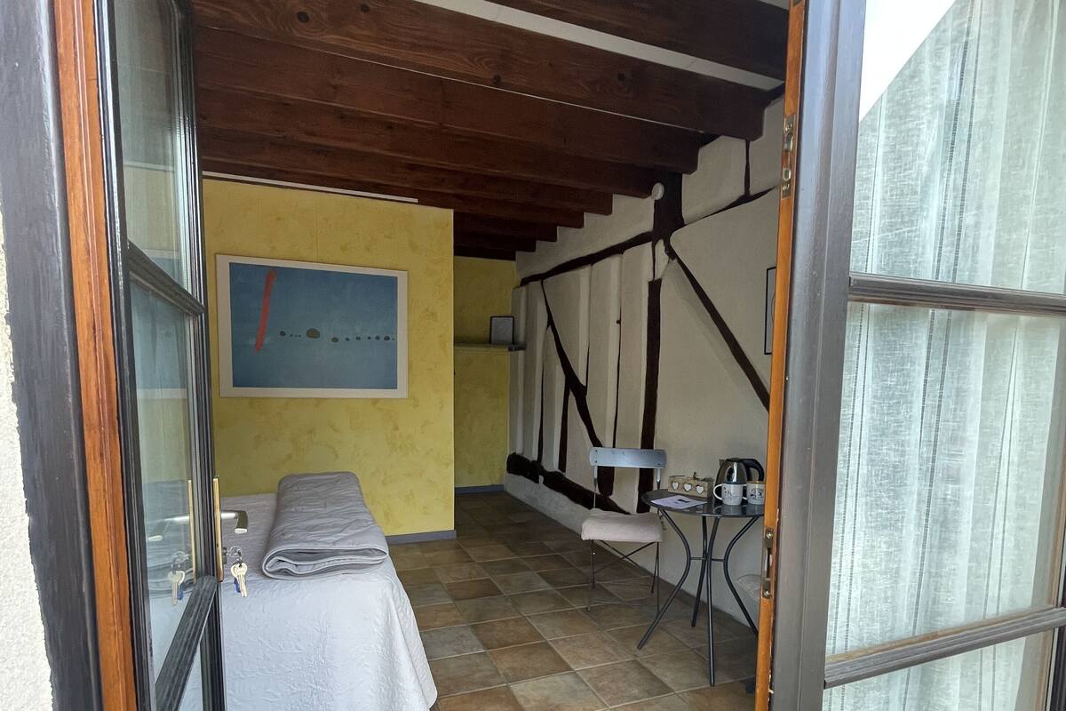 Quarto com cama, uma mesinha e uma cadeira. Há uma janela com cortinas, um quadro na parede e piso de azulejos.