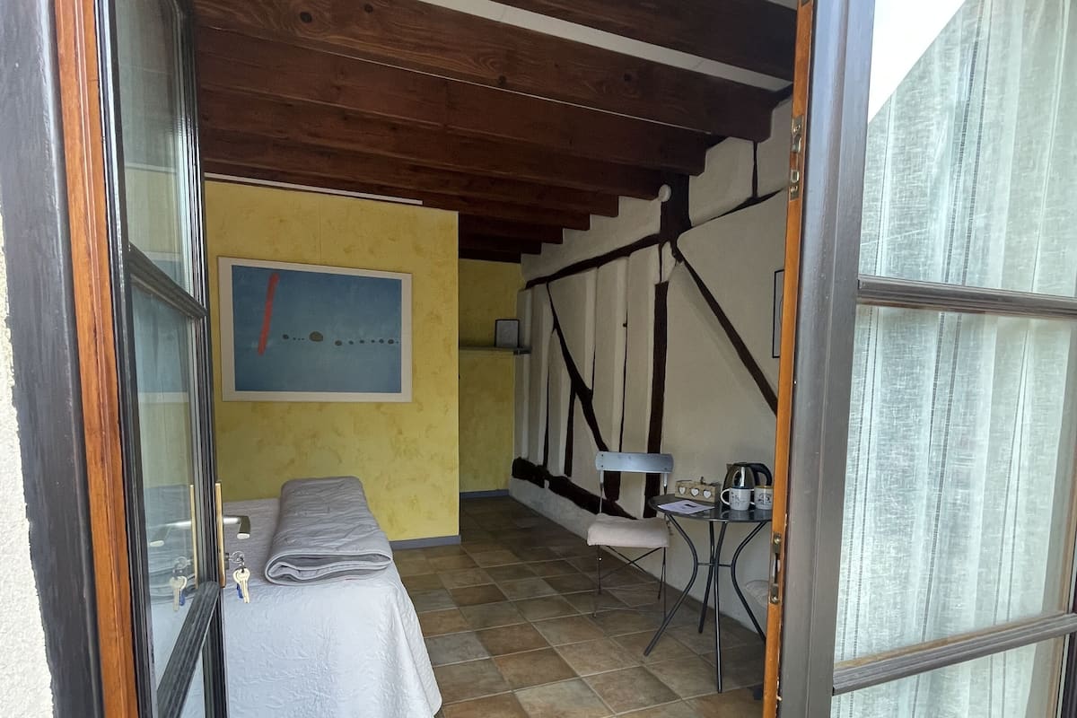 A room with a bed, a small table, and a chair. There is a window with curtains, a painting on the wall, and a tiled floor.