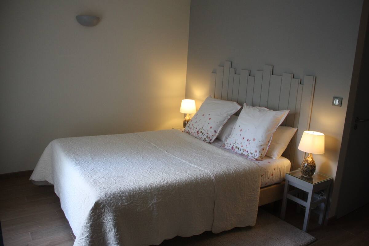Cama bem arrumada com colcha branca e almofadas, duas luminárias de cabeceira e uma mesinha de cabeceira.