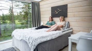 A modern bedroom with a large bed, a wooden wall, a mounted deer antler, and a view of a forest through a window.
