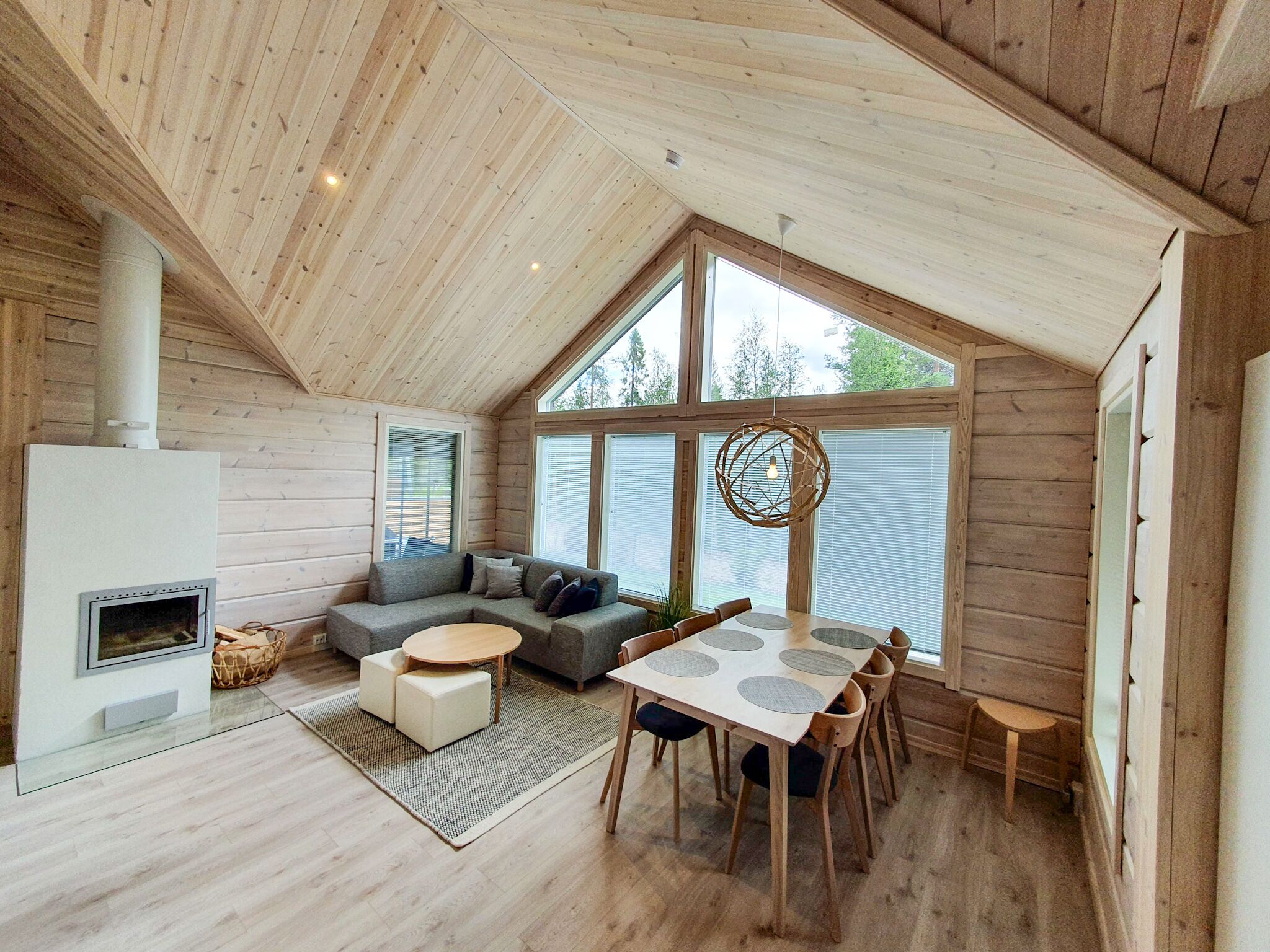 A cozy cabin interior with a loft-style ceiling, a fireplace, a sofa, a dining table, and large windows.