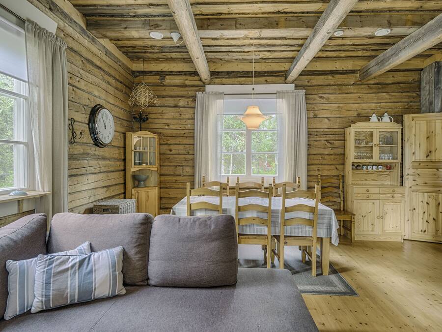 A cozy cabin interior with a wooden ceiling, a dining table with chairs, a sofa with cushions, and wooden cabinets.
