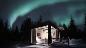A snow-covered landscape with a small, illuminated cabin featuring a visible bedroom and bathroom.