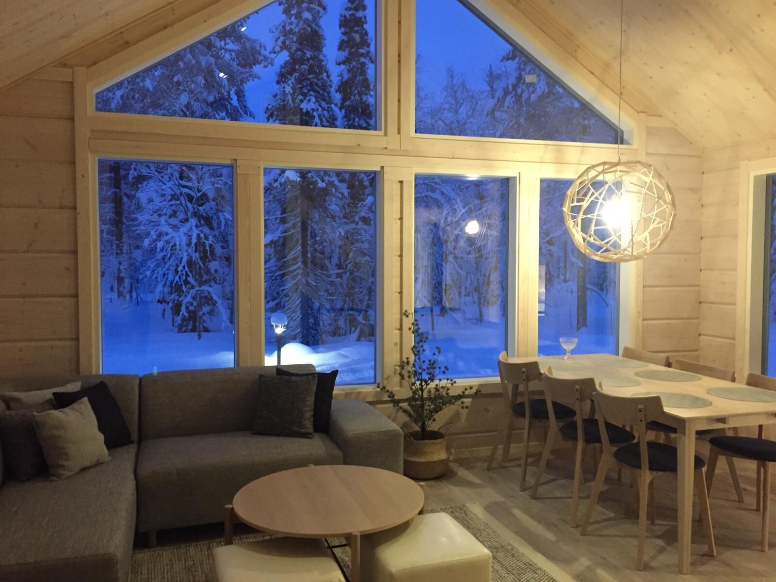 Een gezellig blokhutinterieur met een groot raam dat uitkijkt op een besneeuwd bos, een houten eettafel en een comfortabele zithoek.