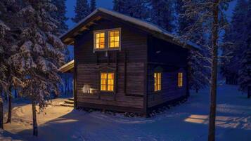 Una cabaña de madera con dos ventanas visibles, rodeada de árboles cubiertos de nieve.