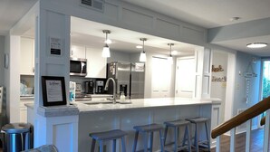 A modern kitchen with a central island, bar stools, stainless steel appliances, and a welcoming sign.