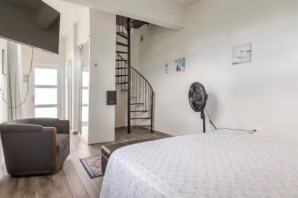 A bedroom with a bed, a chair, a fan, a staircase, and a door leading to another room.