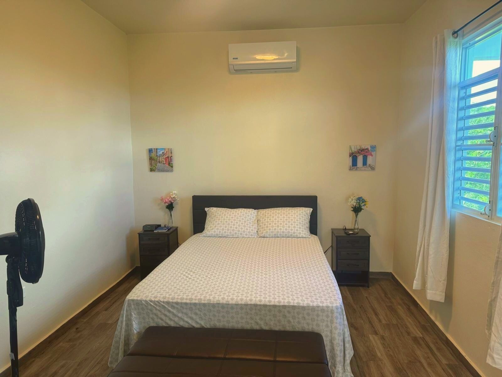Ein Schlafzimmer mit einem Bett, zwei Nachttischen, einem Standventilator und einem Fenster mit Jalousien.