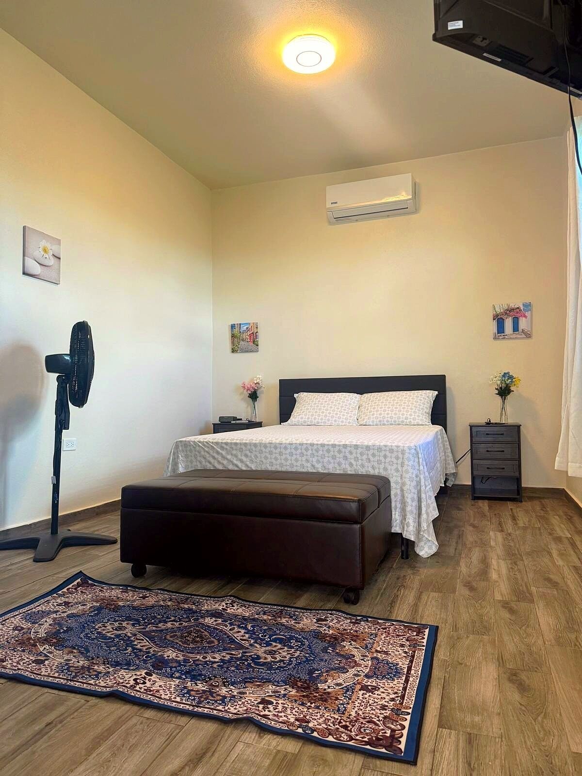 Une chambre à coucher comprenant un lit, des tables de chevet, un ventilateur sur pied, un climatiseur, un tapis et une petite commode.
