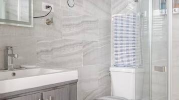Un baño con suelo de mármol, inodoro blanco, cabina de ducha de vidrio y tocador con espejo y lavabo.