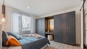Quarto moderno com cama grande, guarda-roupa cinzento e janela com vista para árvores.