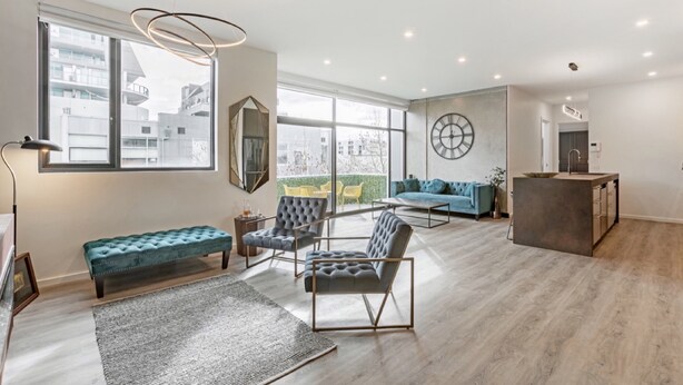 Uma sala de estar ampla, com uma grande janela, um sofá azul, uma poltrona cinza e uma mesa de centro de madeira.