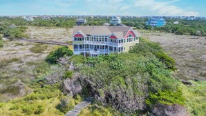 A large, multi-story house with a prominent balcony, surrounded by dense vegetation and a clear view of the ocean. - Soundfront with Private Pool, Hot Tub, Elevator, Private Sound Access, Game Room (Avon)