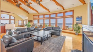 A spacious living room with a large sectional sofa, a coffee table, and a view of the ocean through large windows. - Soundfront with Private Pool, Hot Tub, Elevator, Private Sound Access, Game Room (Avon)