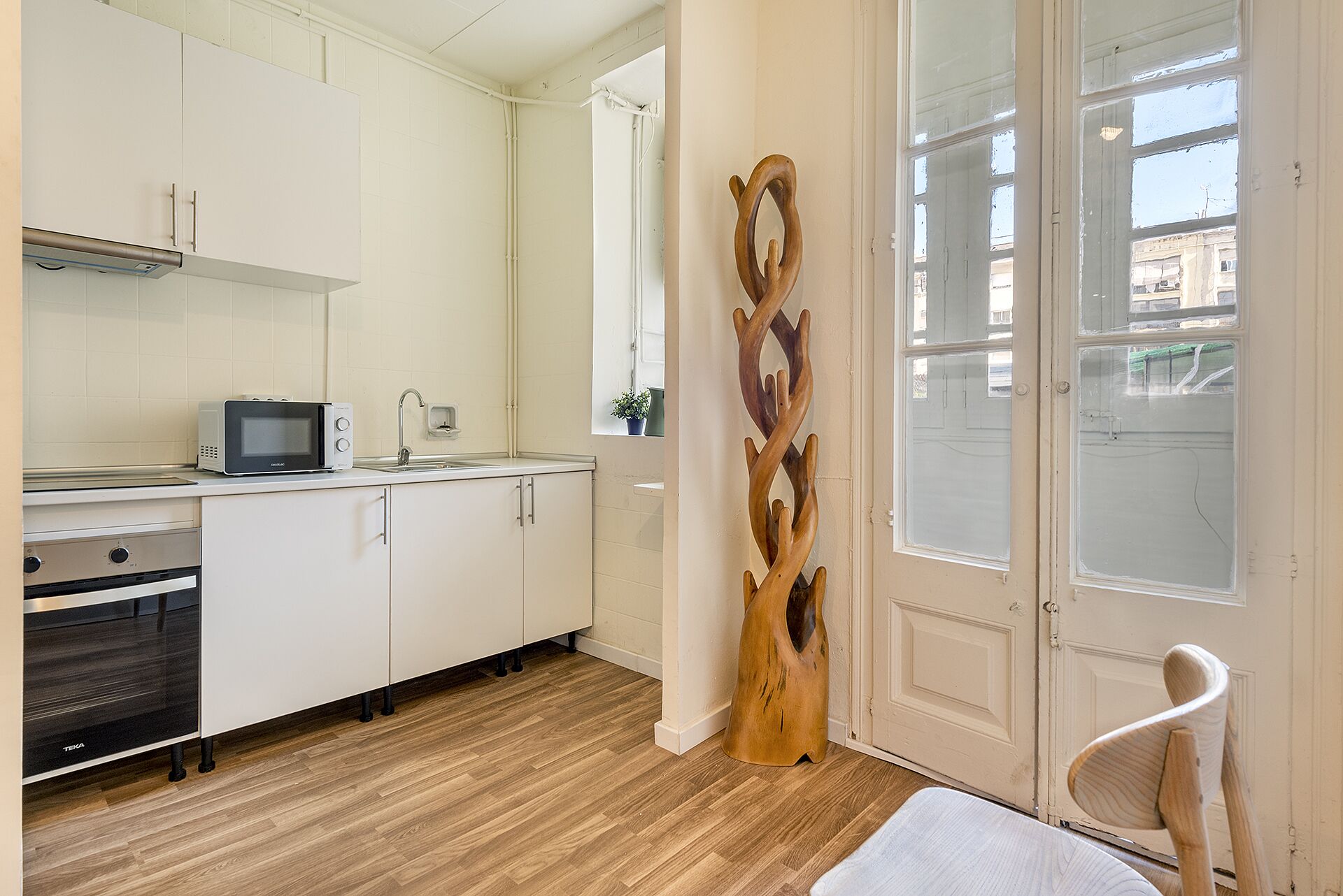 Een moderne keuken met witte kasten, een magnetron en een spoelbak. Er hangt een houten sculptuur aan de muur en er staat een stoel bij de deur.