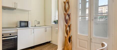 Een moderne keuken met witte kasten, een magnetron en een spoelbak. Er hangt een houten sculptuur aan de muur en er staat een stoel bij de deur.