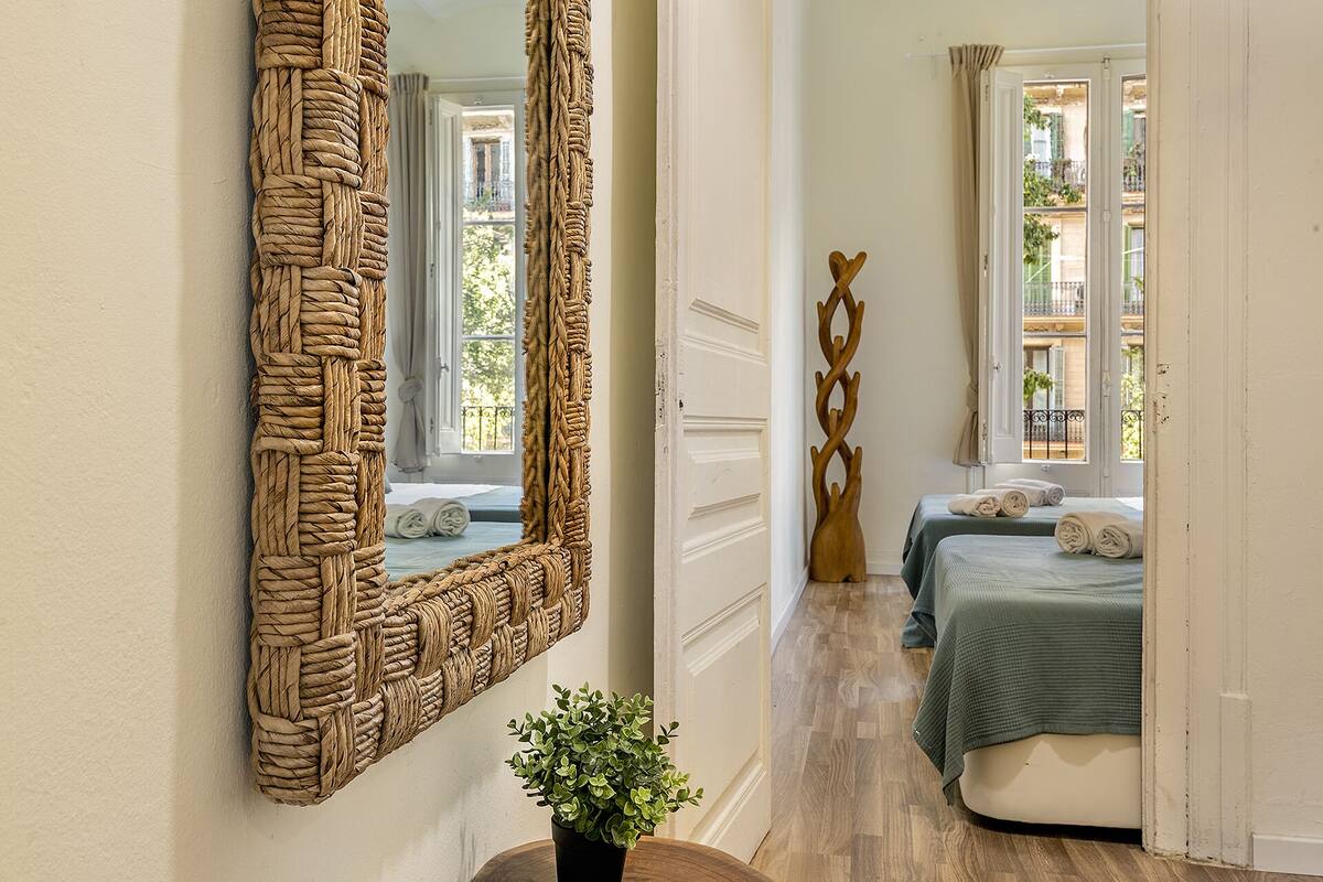 Un pasillo que conduce a un dormitorio con un espejo grande tejido, una escultura de madera y vistas a un balcón.