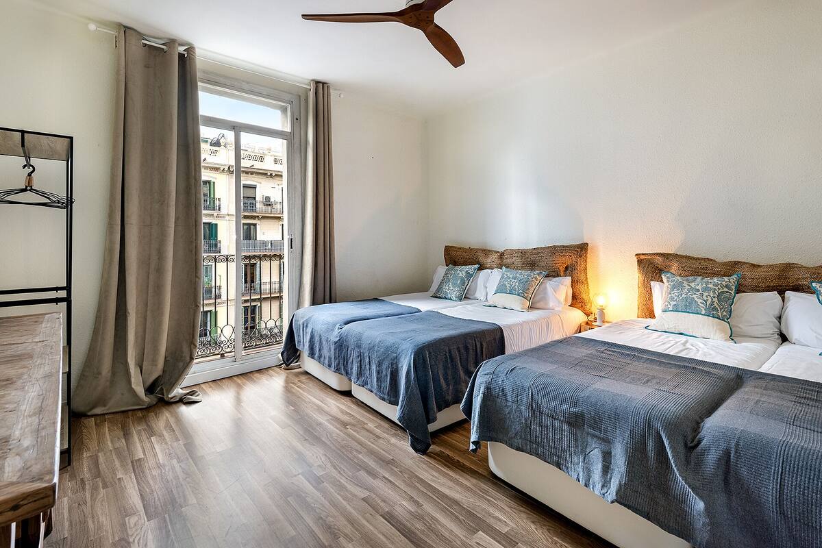 Una habitación de hotel con dos camas, suelo de madera, balcón con cortinas y ventilador de techo.