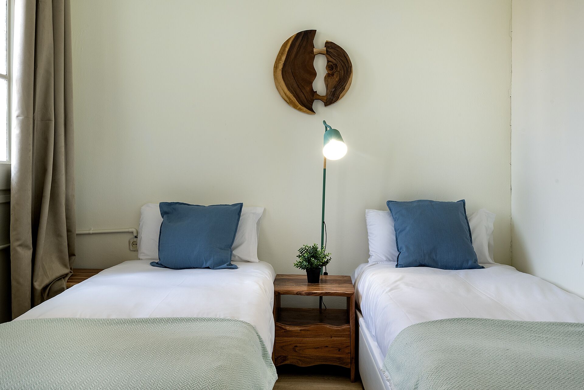 A bedroom with two beds, each with a blue pillow and a green throw blanket. There is a wooden nightstand between the beds with a small potted plant on it. A wall-mounted light fixture is above the nightstand.