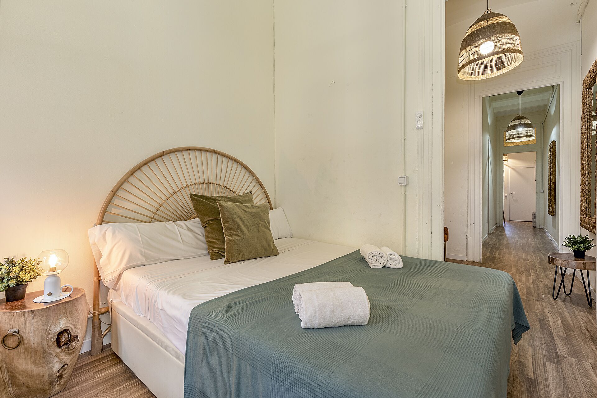A bedroom with a round headboard, a bed with white linens, a green throw blanket, and a white towel. There is a bedside table with a lamp and a potted plant.