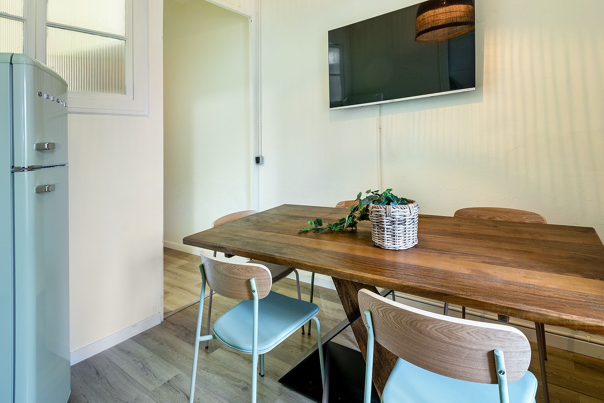 Un comedor con una mesa de madera, dos sillas, una cesta con follaje y un televisor montado en la pared.