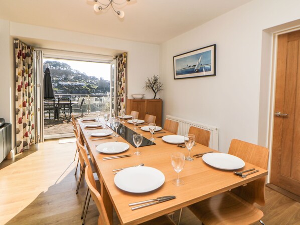 A dining area with a long wooden table set for six, a view of a balcony, and a framed picture on the wall.
