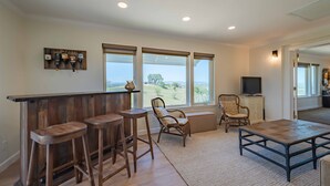 A spacious living area with a wooden bar, two cushioned chairs, a television, and large windows offering a view of the outdoors. - Hilltop Vineyard Estate with 360 Views, Hot Tub & Pool! (Paso Robles)