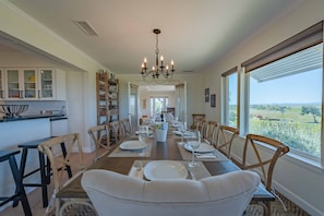 Uma sala de jantar com uma mesa comprida posta para seis pessoas, ladeada por cadeiras de madeira, um candelabro e grandes janelas com vista para o campo.