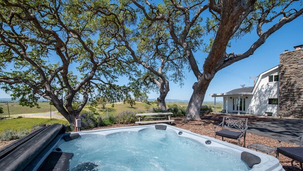 A hot tub with a stone exterior, surrounded by a patio area with outdoor furniture, a stone wall, and a large tree. - Hilltop Vineyard Estate with 360 Views, Hot Tub & Pool! (Paso Robles)