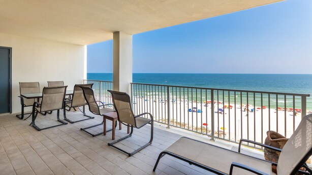 Varanda com quatro cadeiras e uma mesa, com vista para uma praia com guarda-sóis e pessoas.