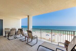 Un balcon avec quatre chaises et une table, donnant sur une plage parsemée de parasols et où se trouvent des gens.