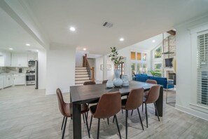 Una cocina moderna con armarios blancos, un comedor con mesa de madera oscura y sillas marrones, y una escalera que lleva a una sala de estar con un sofá azul y una chimenea.