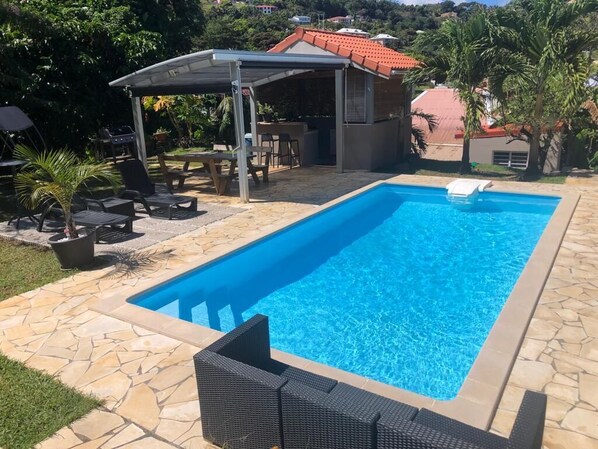 A rectangular swimming pool with a stone border, surrounded by lounge chairs and a covered patio area. - A 10mn DE LA Plage Evasion Créole Villa Entre Nature & Modernité (La Trinité)