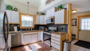 Une cuisine avec des armoires blanches, un réfrigérateur en acier inoxydable, un four à micro-ondes et une cuisinière. Il y a une fenêtre avec un rideau, un tapis sur le sol et une poubelle.