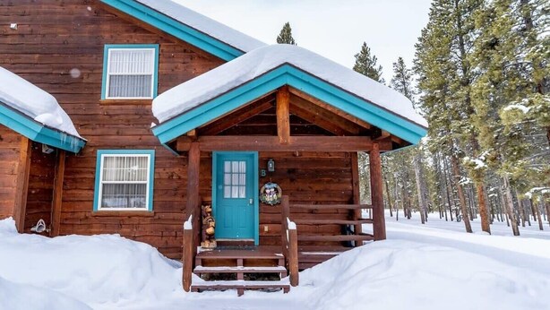 Une cabane en bois avec une porte et des fenêtres bleues, entourée d’un paysage enneigé et d’arbres.