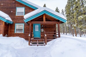A wooden cabin with a blue door and windows, surrounded by snow-covered ground and trees.