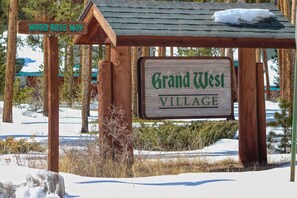 Ein hölzernes Wegweiser mit der Aufschrift 'Grand West Village', umgeben von Schnee und Bäumen.