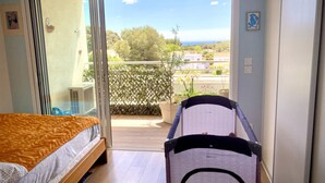 A bedroom with a bed, a changing table, and a view of a balcony with plants and a distant landscape. - Appartement T2 Vue Mer (Martigues)