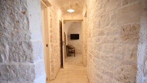 Ein schmaler Korridor mit Steinwänden und Fliesenboden, der zu einem Raum mit an der Wand montiertem Flachbildfernseher führt.
