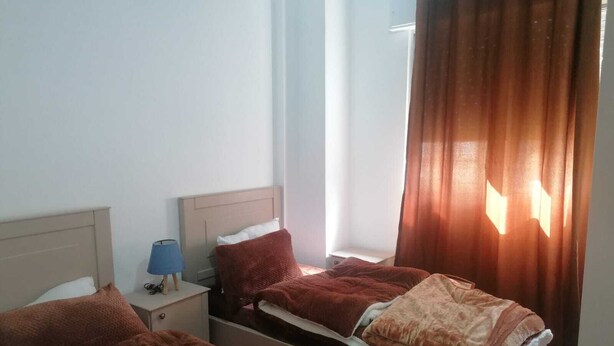 A hotel room with two beds, a nightstand with a lamp, and a window with curtains. - Furnisher apartment in Al Jubiha (Amman)