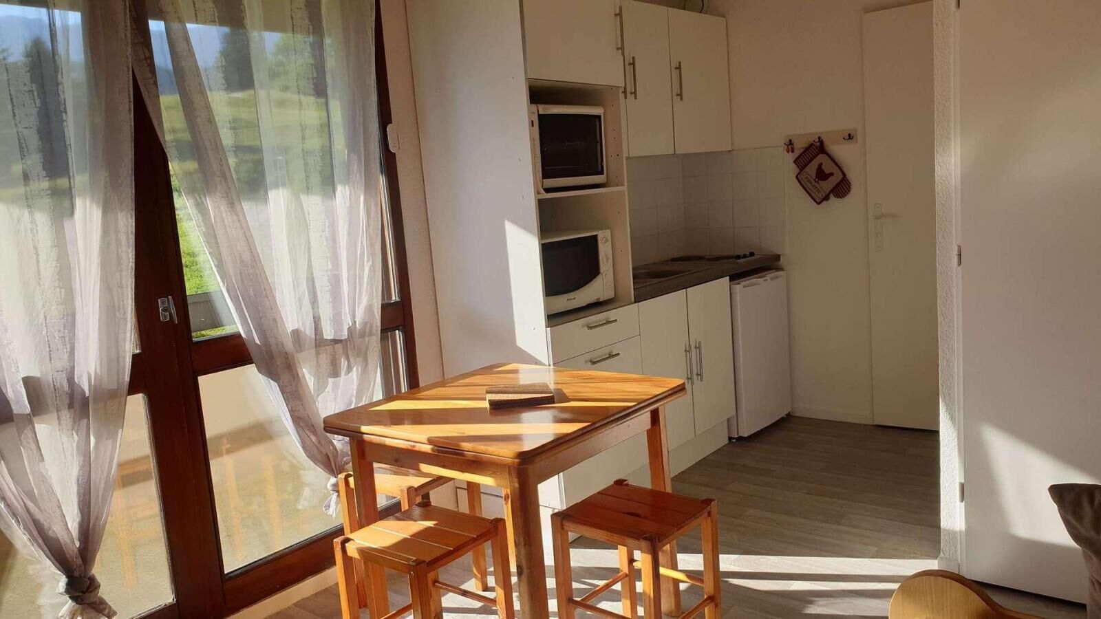 Eine kleine Küche mit weißen Schränken, einer Mikrowelle und einem Geschirrspüler. Ein Holztisch mit Stühlen und ein Fenster mit durchsichtigen Vorhängen.