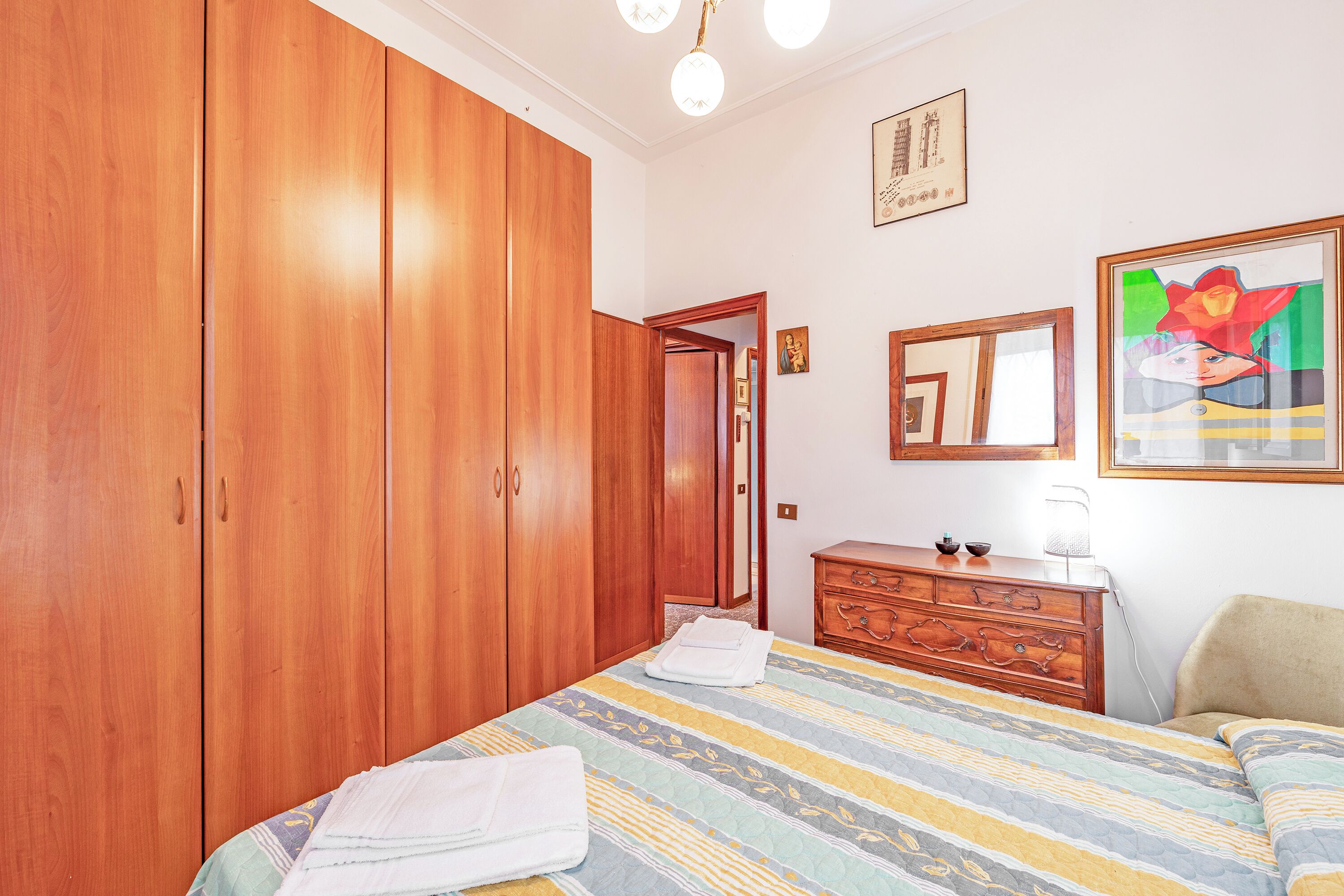A bedroom with a large wardrobe, a bed with a striped bedspread, a dresser, and a framed picture on the wall.