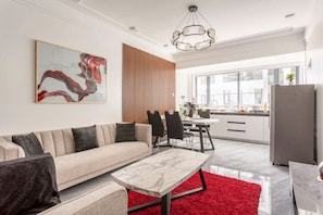 A modern living room with a sofa, a marble coffee table, and a red rug. There is a dining area with a table and chairs, and a kitchen with a refrigerator. A large window provides a view of the city. - Inikas Living_Luxurious 1 bedroom apartment  (Nairobi)